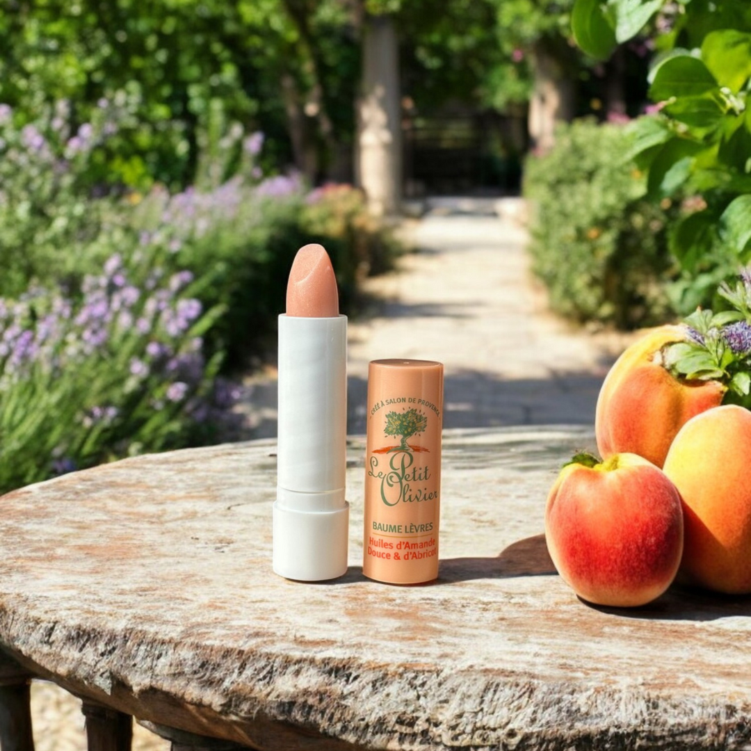 An open tube of Baume Lèvres Soin & Brillance sits on a round wooden table outdoors beside fresh peaches, with a sunlit garden path, green trees, and purple flowers in the background.