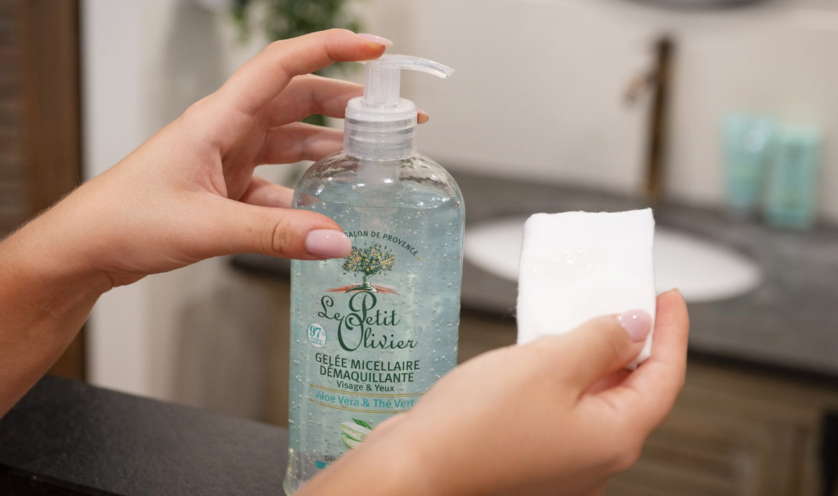Une personne verse du démaquillant en gel de la marque « Le Petit Olivier » sur un coton, avec en arrière-plan un lavabo de salle de bains et un décor flou.
