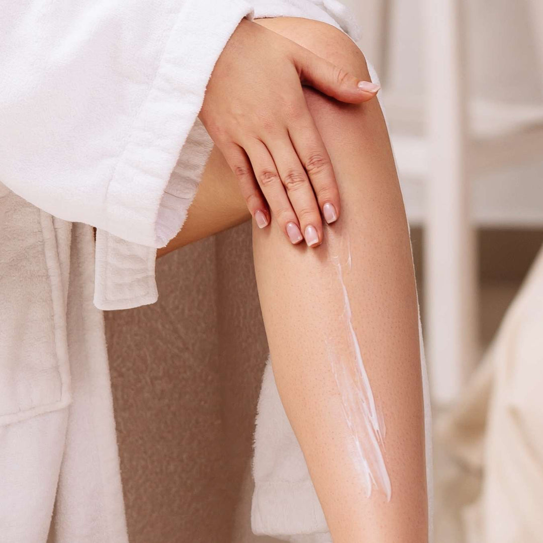 Une personne vêtue d'une robe blanche applique une lotion ou une crème sur sa jambe, en l'étalant délicatement avec la main.