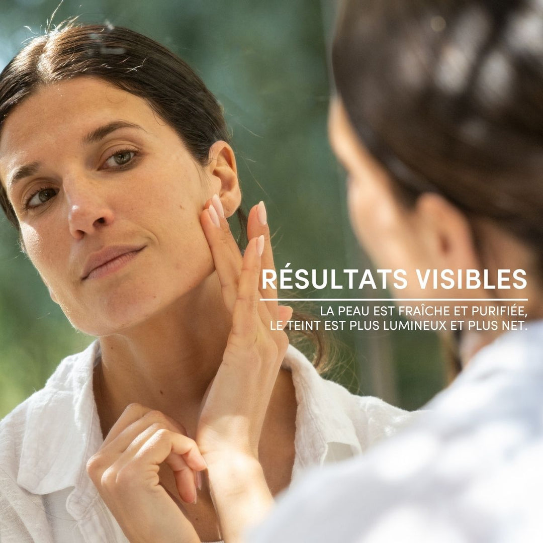 Une femme aux cheveux foncés se touche le visage dans une pièce baignée d'une lumière tamisée, vêtue d'un haut blanc. Le texte en français indique « RÉSULTATS VISIBLES », mettant en avant une peau plus nette et éclatante grâce au Masque Visage Stick Pureté de Le Petit Olivier.