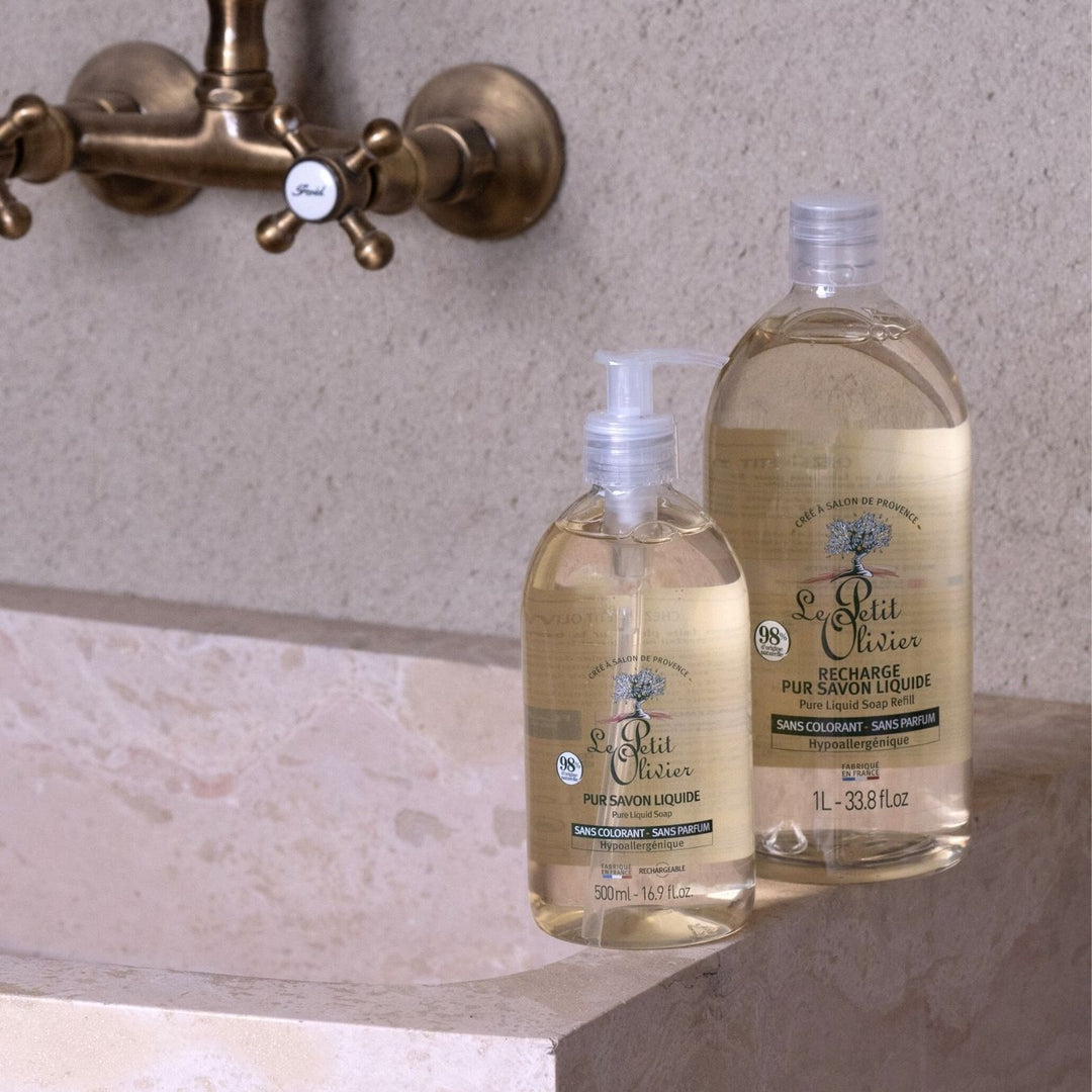 Deux flacons de « Le Petit Olivier Recharge Pur Savon Liquide - Sans Parfum », l'un de 500 ml et l'autre de 1 litre, reposent sur un lavabo en pierre beige, sous un robinet vintage en laiton. Le logo représentant un arbre et la mention « base végétale » ressortent sur les étiquettes.