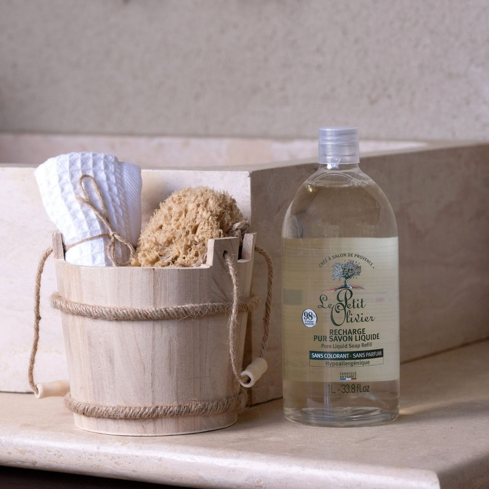 Un seau en bois muni d'une poignée en corde contient une serviette blanche et une éponge naturelle, posés à côté d'un flacon de « Le Petit Olivier Recharge Pur Savon Liquide - Sans Parfum » sur un plan de travail beige, avec un lavabo en arrière-plan.