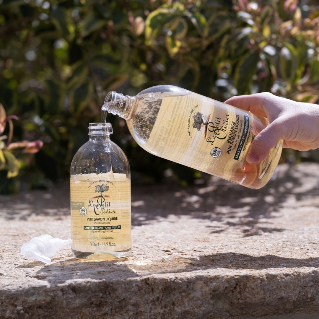 Une main verse du savon liquide « Le Petit Olivier Recharge Pur » – sans parfum, à base végétale – d’un grand flacon dans un plus petit, à l’extérieur, sur une surface en pierre entourée d’une végétation luxuriante.