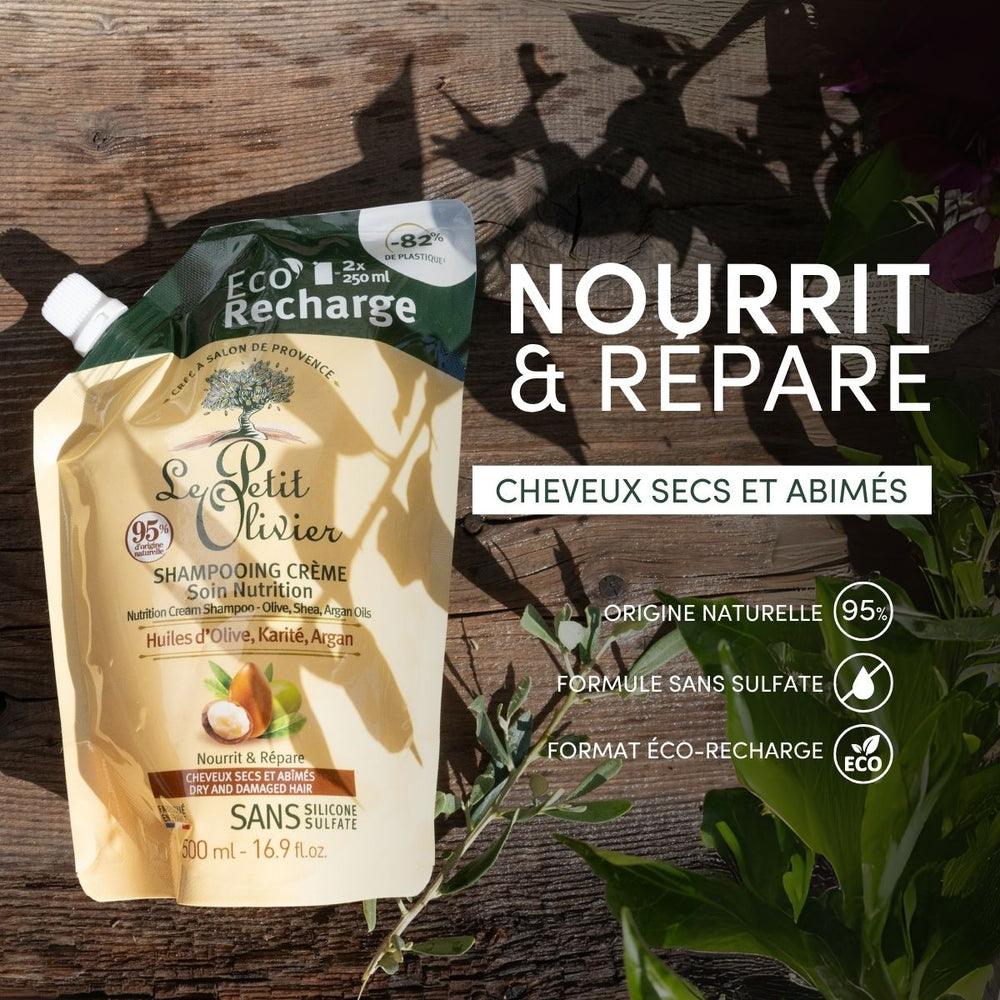 Un sachet de « Le Petit Olivier Eco-Recharge – Shampooing pour cheveux secs et abîmés » repose sur une surface en bois, baignée par la lumière du soleil et les ombres des plantes. L'emballage de recharge écologique met en avant les huiles d'olive, de karité et d'argan, ainsi qu'une formule sans sulfate.