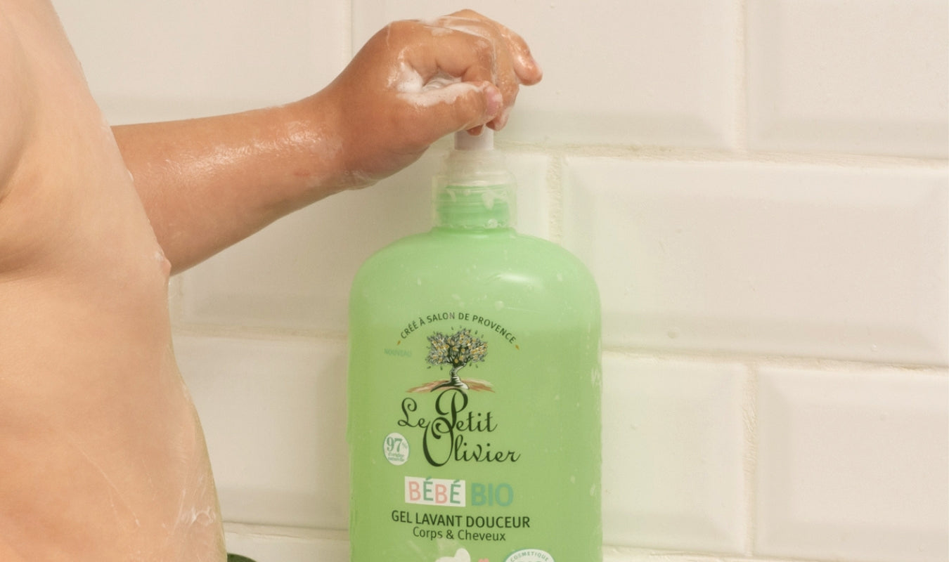 La main savonneuse d'un enfant appuie sur la pompe d'un flacon vert de gel douche et shampoing doux « Le Petit Olivier Bébé Bio », adossé à un mur carrelé blanc.