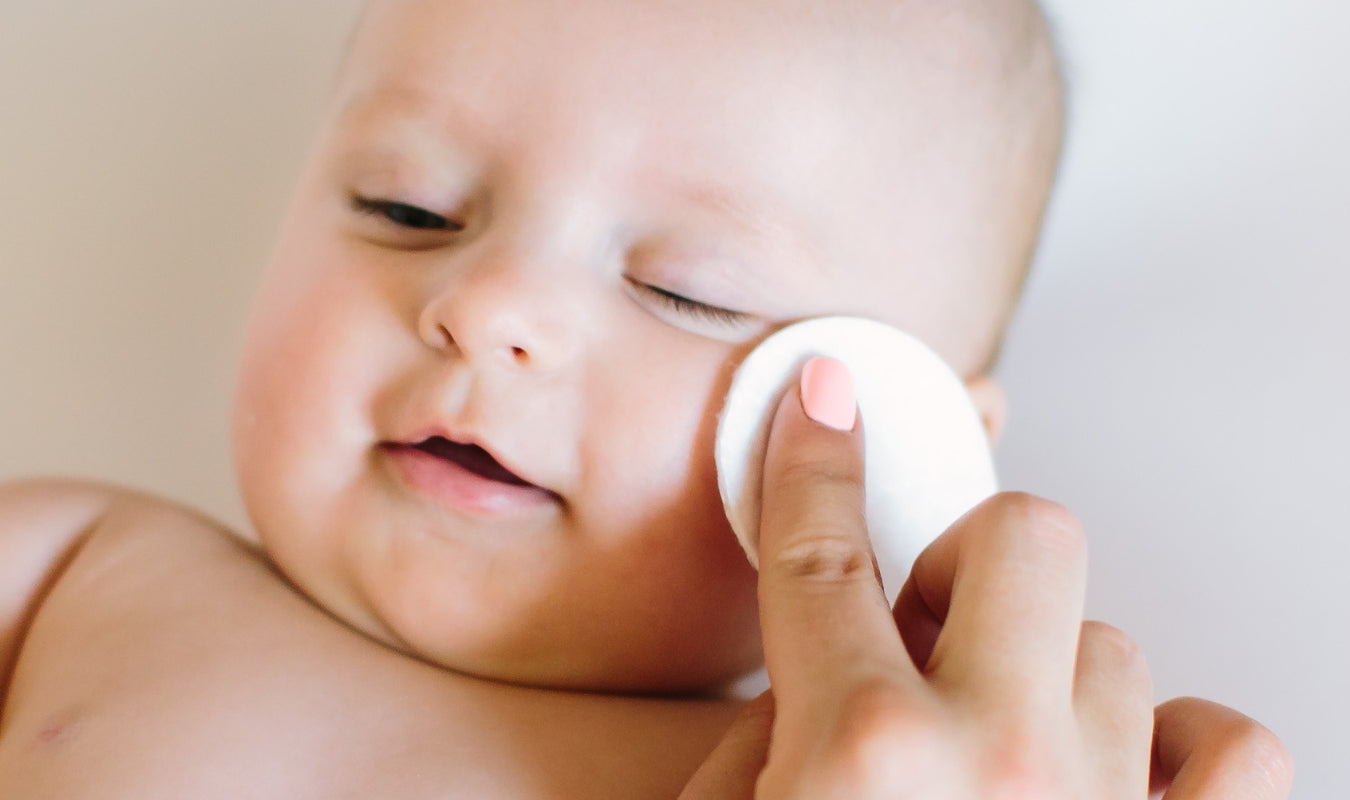 Un adulte aux ongles vernis d'un rose pâle essuie délicatement la joue d'un bébé à l'aide d'un coton.
