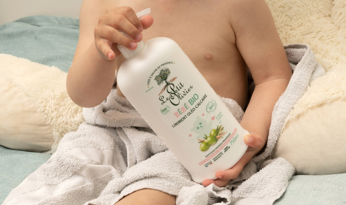 Un petit enfant enveloppé dans une serviette tient une grande bouteille blanche de lotion nettoyante « Le Château Le Chat Bébé Bio », dont l'étiquette est ornée d'une illustration représentant une olive, assis sur un lit recouvert d'une literie aux couleurs claires.