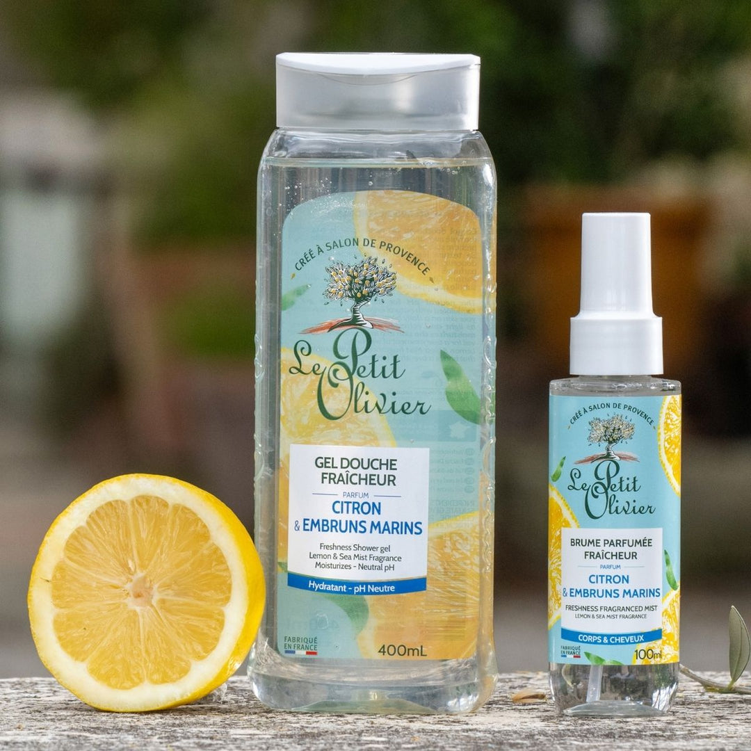 Deux produits de la marque Le Petit Olivier — la « Brume Parfumée Fraîcheur » et le gel douche, tous deux parfumés au citron et aux embruns marins — sont présentés en extérieur, à côté d'un citron coupé en deux. Leurs étiquettes bleues et jaunes arborent des motifs de citron, avec une végétation légèrement floue en arrière-plan.