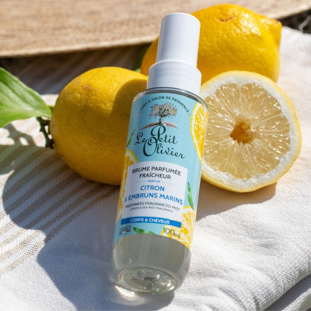 Un flacon de « Le Petit Olivier Brume Parfumée Fraîcheur » repose sur un tissu rayé, à côté de deux citrons entiers et d'un citron coupé en deux, tandis que la lumière du soleil projette de douces ombres sur la scène.