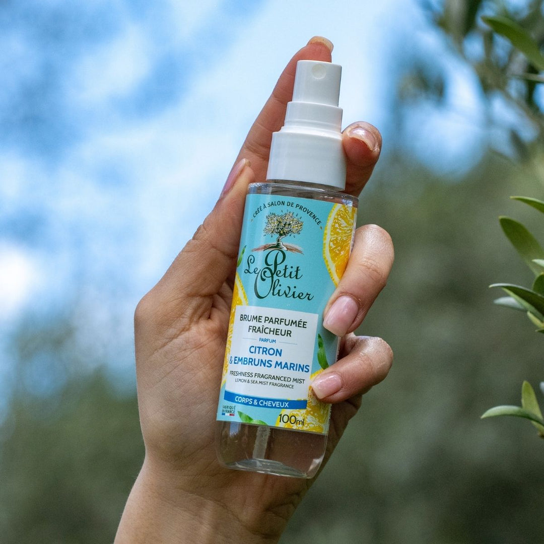 Une main tient le vaporisateur « Le Petit Olivier Brume Parfumée Fraîcheur », aux notes de citron et d'océan, idéal pour rafraîchir les cheveux, sur fond flou d'un paysage verdoyant en plein air.