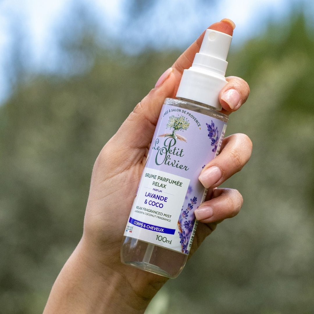 Une main tient un flacon de brume parfumée pour le corps « Le Petit Olivier Brume Parfumée Relax », conçue pour le corps et les cheveux, avec en arrière-plan une végétation floue en plein air.