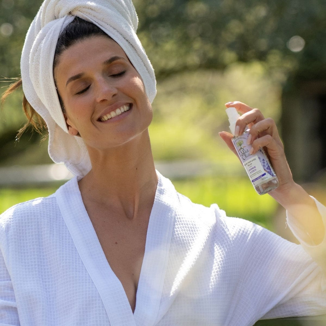 Une femme vêtue d'un peignoir blanc et d'un turban de serviette sourit, les yeux fermés, tout en vaporisant la Brume Parfumée Relax de Le Petit Olivier pour le corps et les cheveux, en plein air, au milieu d'une végétation luxuriante.