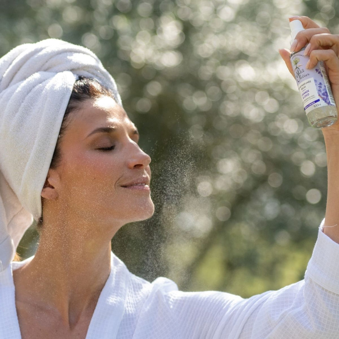 Une femme vêtue d'une robe blanche, une serviette sur la tête, vaporise « Le Petit Olivier Brume Parfumée Relax » sur son visage à l'extérieur, les yeux fermés et le sourire aux lèvres, profitant d'un moment de soin rafraîchissant pour le corps et les cheveux.