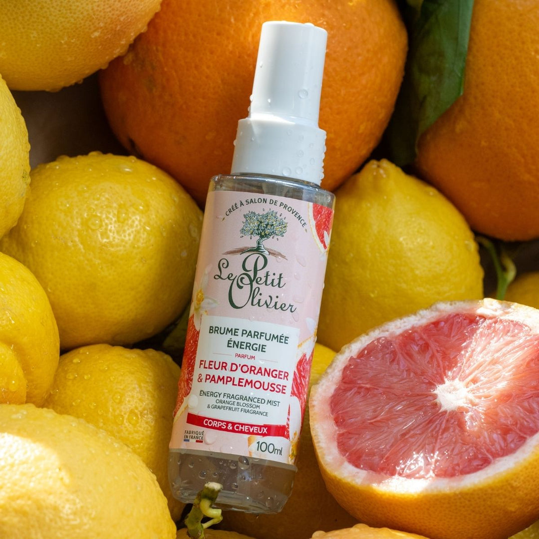 Un flacon de « Le Petit Olivier Brume Parfumée Énergie » trône au milieu de citrons frais, d'oranges et d'un pamplemousse coupé en deux. Le flacon transparent est muni d'un bouchon vaporisateur blanc et d'une étiquette rose.