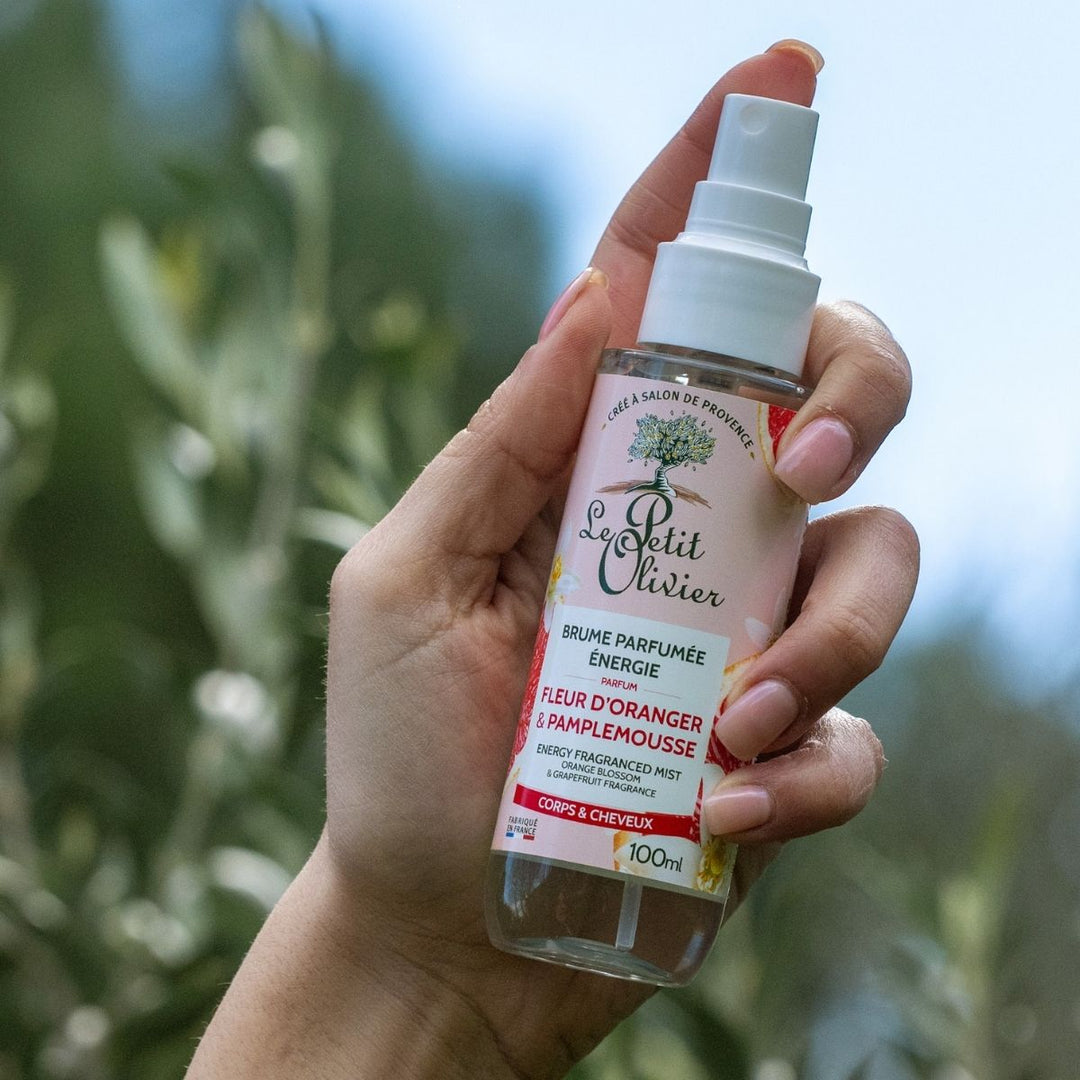 Une main aux ongles soignés tient le flacon de « Le Petit Olivier Brume Parfumée Énergie » sur un fond vert flou. L'étiquette met en avant son parfum rafraîchissant, idéal pour redonner de l'énergie au corps et aux cheveux.