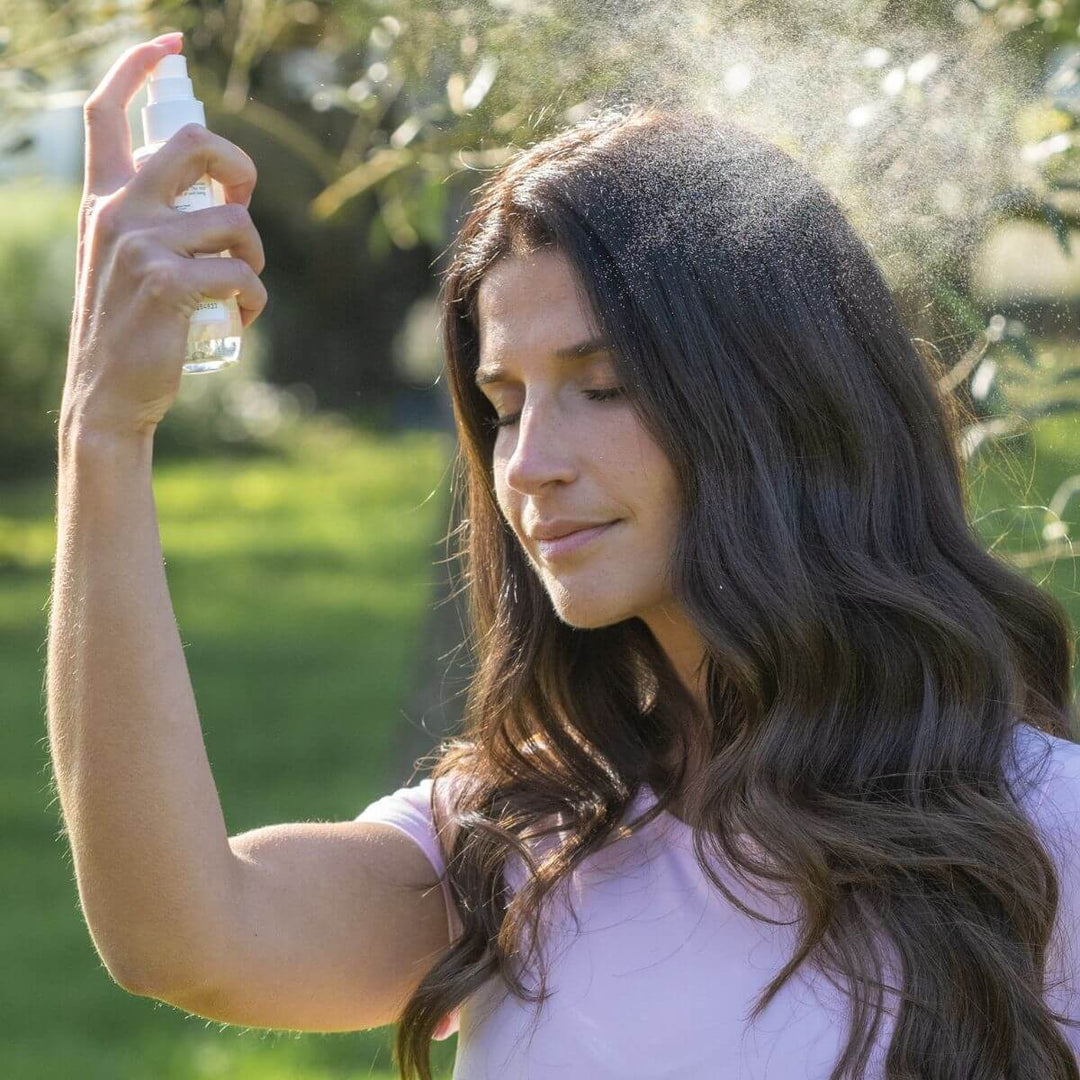 Une femme aux longs cheveux châtains, vêtue d'un t-shirt rose pâle, vaporise la Brume Parfumée Énergie de Le Petit Olivier sur son visage, debout en plein air dans un cadre verdoyant et ensoleillé, l'air détendu.