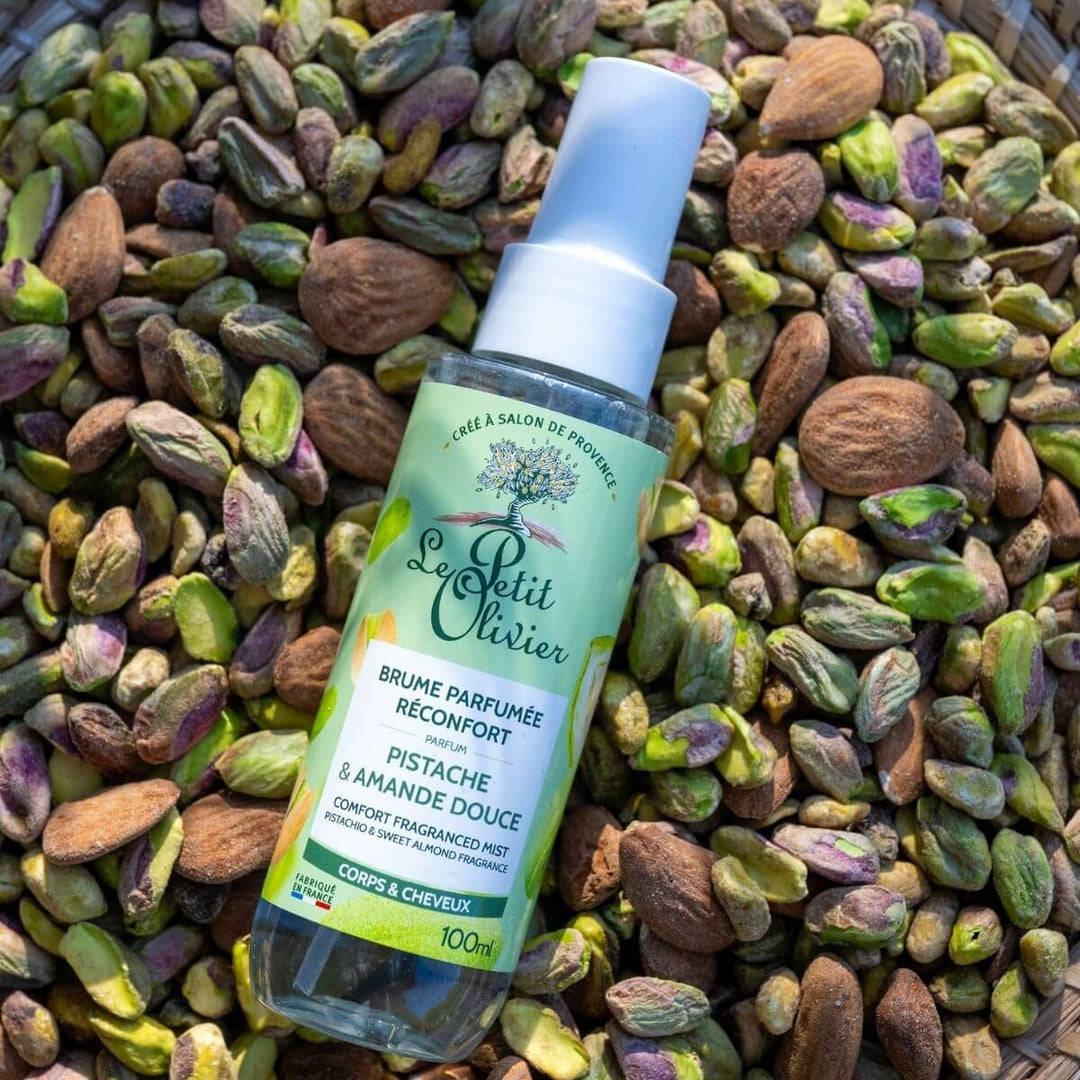Un flacon de « Le Petit Olivier Brume Parfumée Réconfort » repose sur des pistaches entières et décortiquées ; la lumière du soleil illumine à la fois le produit et les fruits à coque, évoquant ainsi ce parfum réconfortant de fabrication française.
