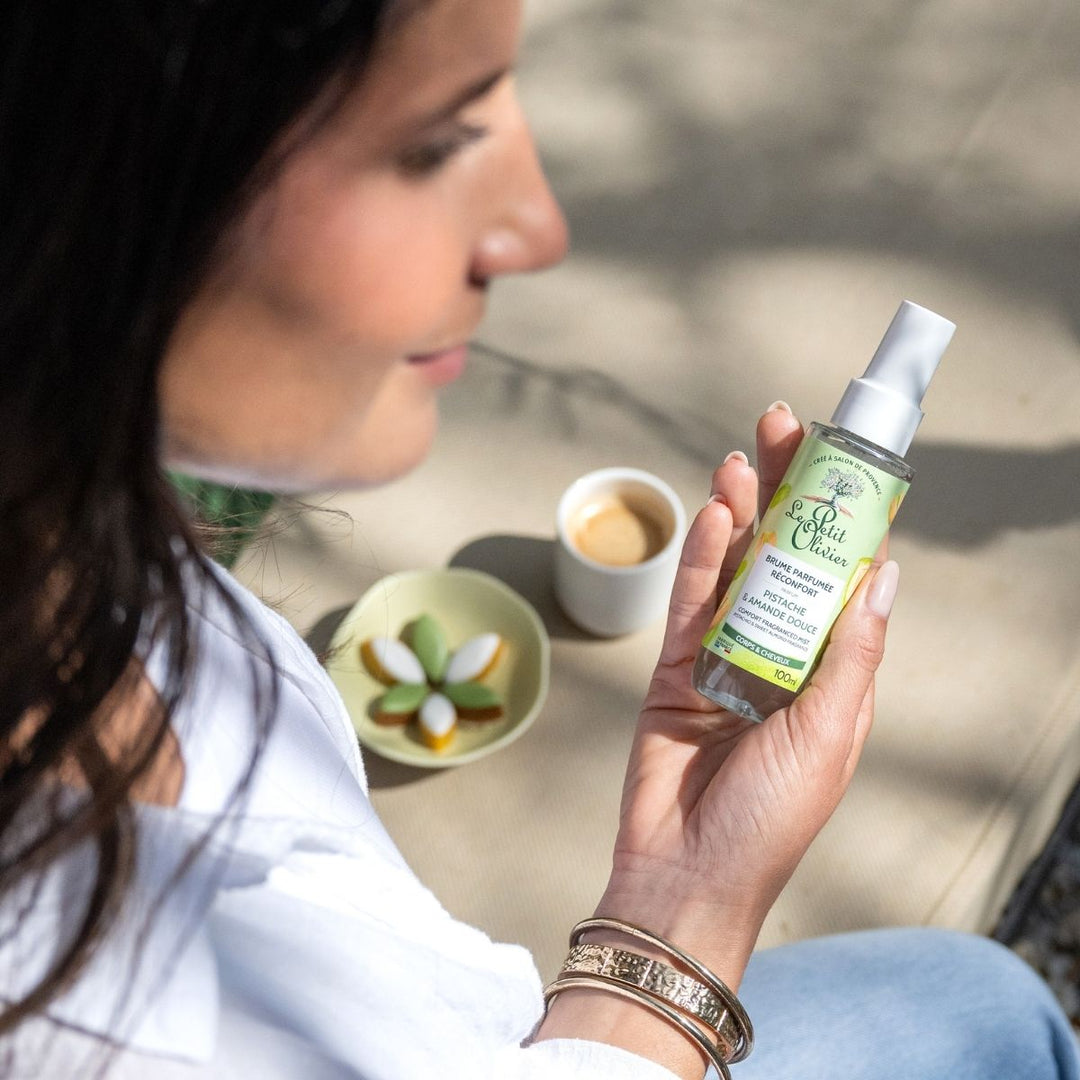 Une femme tient un flacon de « Le Petit Olivier Brume Parfumée Réconfort », fabriqué en France, tout en se détendant en plein air avec un café et des friandises aux couleurs pastel — savourant un moment aussi délicieux que la pistache à l'amande douce.