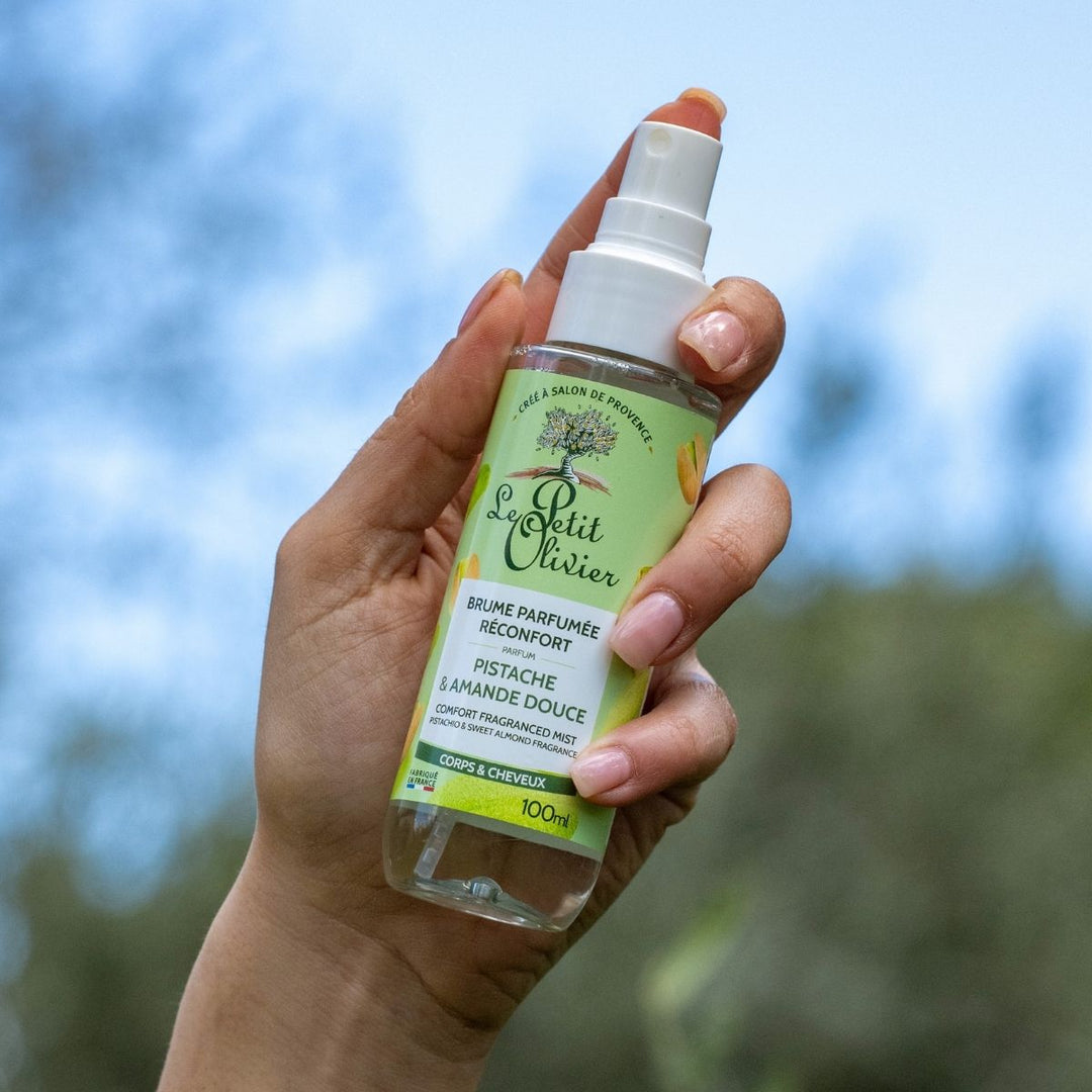 Une main tient un vaporisateur de « Le Petit Olivier Brume Parfumée Réconfort », fabriqué en France, à l'extérieur, sur fond flou de verdure composé d'arbres ou de plantes.
