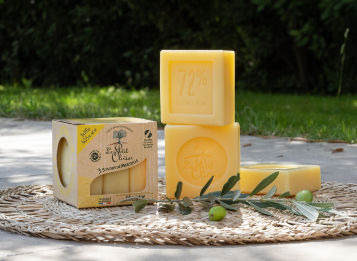 Trois gros pains de savon jaunes et une boîte portant l'inscription « Le Petit Olivier » sont posés sur une natte tressée à l'extérieur, avec des branches d'olivier et des feuilles vertes en arrière-plan.
