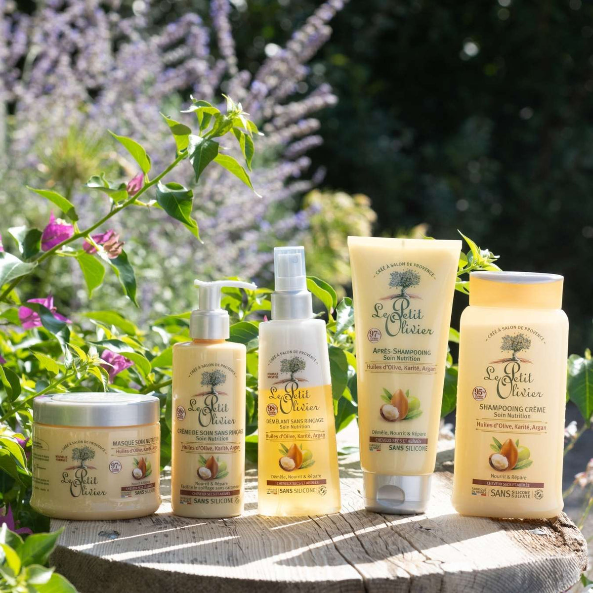 Cinq flacons et pots de produits de soins capillaires « Le Petit Olivier » sont disposés à l'extérieur sur une surface en pierre, entourés de plantes vertes et de fleurs violettes, sous un soleil éclatant.