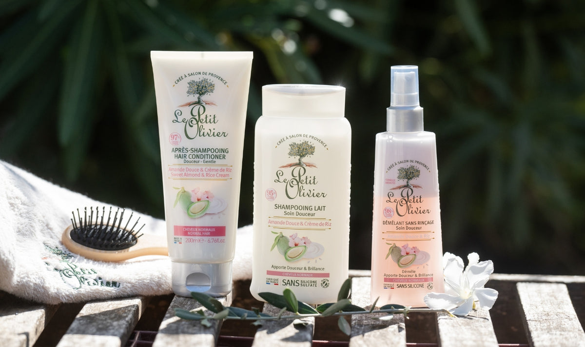 Trois produits de soin capillaire « Le Petit Olivier » — un après-shampoing, un shampoing et un spray démêlant — sont présentés sur un support en bois, accompagnés d'une brosse, d'une serviette blanche et de fleurs blanches, sur fond flou représentant un décor extérieur verdoyant.
