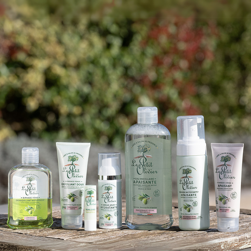 Six produits de la gamme « Le Petit Olivier Rituel Soin Visage Complet Douceur », idéaux pour les peaux normales à sèches et dont l'ingrédient phare est l'olive, sont présentés en rangée à l'extérieur sur un support en bois, avec une végétation floue en arrière-plan.