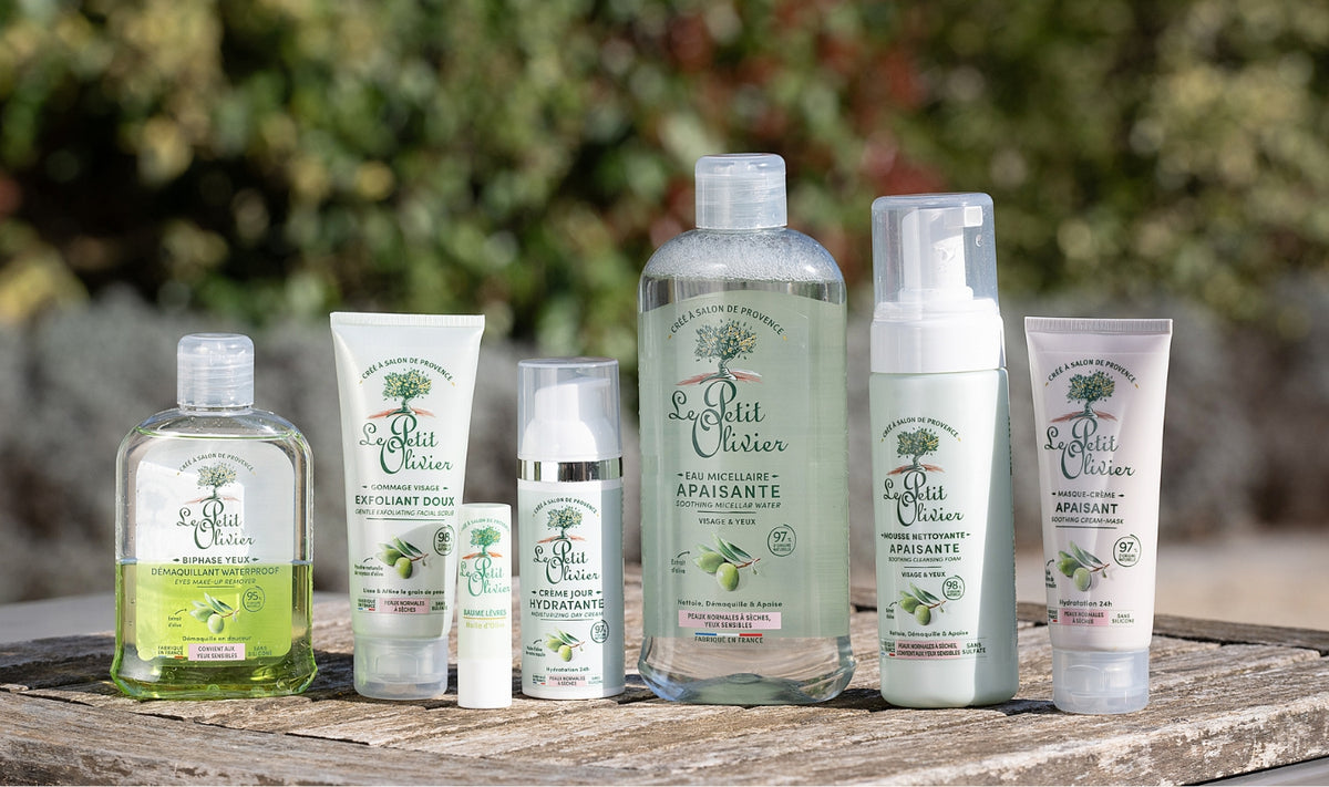 Six produits de soin « Le Petit Olivier » sont disposés sur une surface en bois à l'extérieur, notamment de l'eau micellaire, un nettoyant et une crème hydratante, tous présentés dans des flacons aux emballages verts, blancs et transparents. On aperçoit un arrière-plan feuillu et flou.