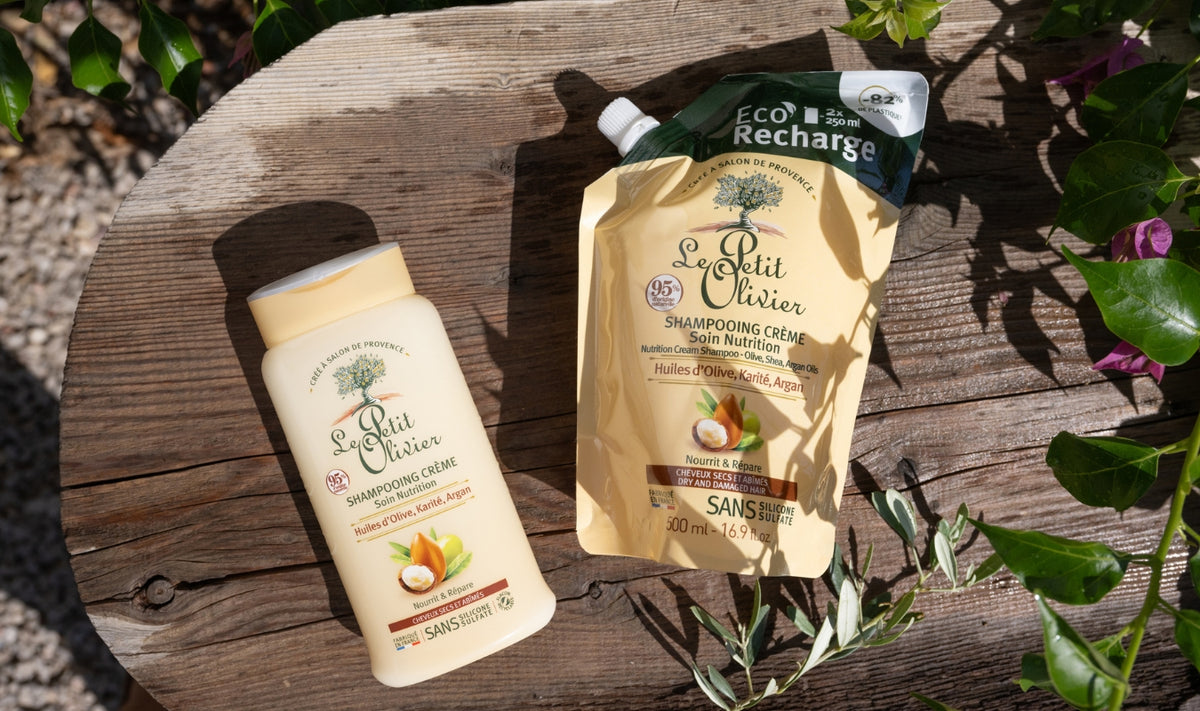 Deux flacons de shampoing « Le Petit Olivier » aux huiles d'argan, d'olive et de karité sont exposés à l'extérieur, au milieu des plantes. L'un est un flacon en plastique ; l'autre est un sachet de recharge écologique.