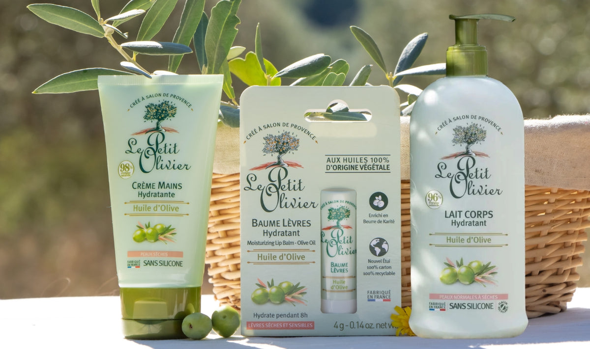 Trois produits de soin « Le Petit Olivier » à l'huile d'olive sont présentés en extérieur sur un fond blanc, avec un panier en osier et des branches d'olivier en arrière-plan. Il s'agit d'une crème pour les mains, d'un baume à lèvres et d'une lotion pour le corps.