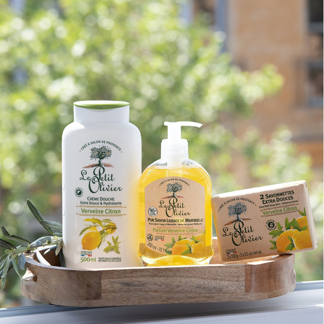 Trio Vivifiant - Douche & Savon - Parfum Verveine-Citron: a set of Le Petit Olivier bath products—Extra Gentle Cream, liquid soap, and Savon de Marseille bar soaps—made in France and beautifully displayed outdoors among green foliage.