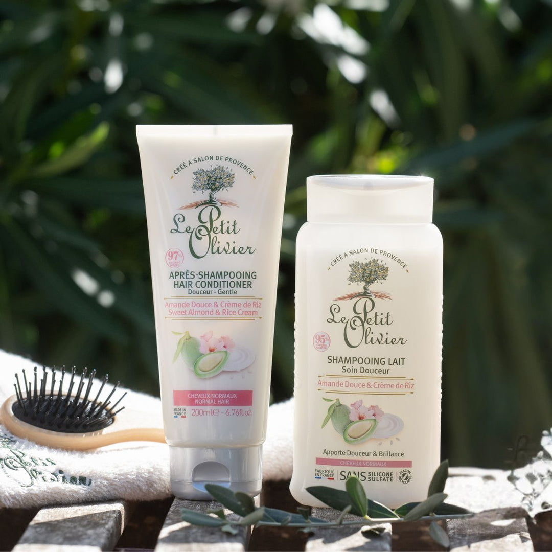 Deux flacons de « Le Petit Olivier Duo Shampooing et Après-shampooing - Cheveux Normaux » sont posés sur une surface en bois, à côté d'une brosse à cheveux, avec de la verdure et des rayons de soleil en arrière-plan.