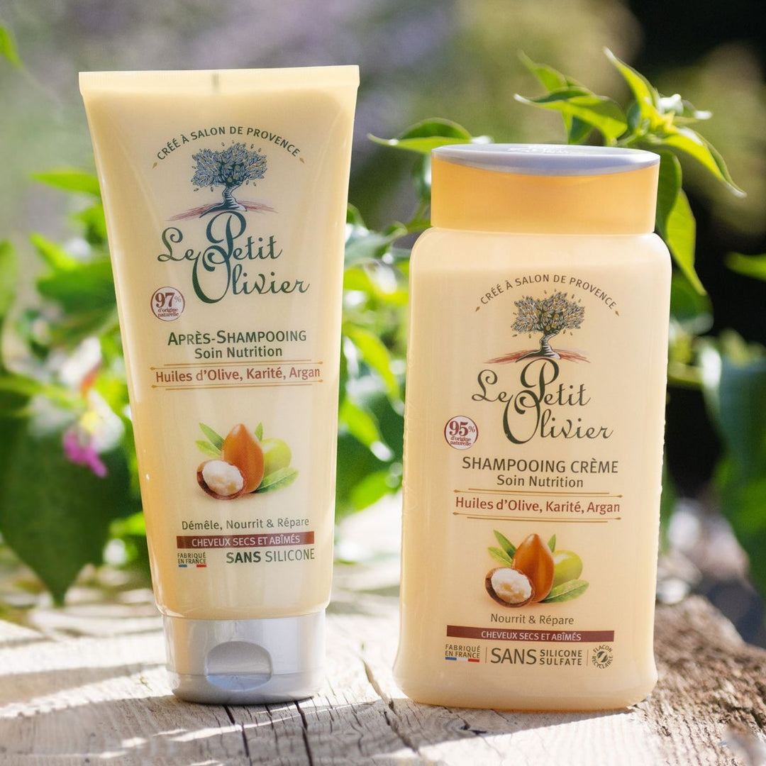 Deux flacons jaunes de « Le Petit Olivier Duo Shampooing et Après-shampooing – Cheveux secs et abîmés » reposent à l'extérieur sur du bois ; enrichis en huiles d'olive, de karité et d'argan, ils sont entourés de verdure et baignés par la lumière du soleil.
