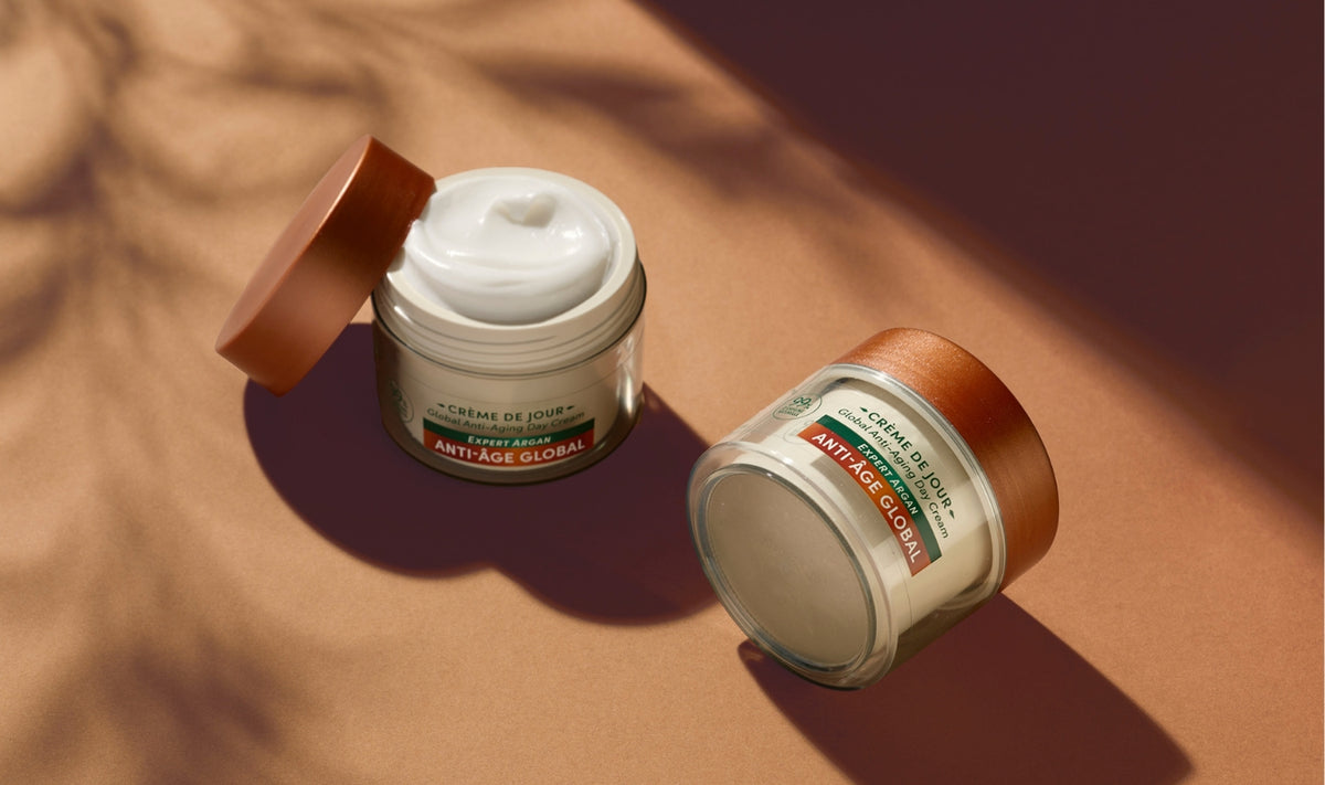 Deux pots de crème anti-âge pour le visage, dotés de couvercles marron, sont posés sur une surface marron clair. L'un des pots est ouvert, laissant entrevoir la crème blanche qu'il contient, tandis que l'autre est fermé. Les ombres et la lumière du soleil créent une ambiance chaleureuse et naturelle.