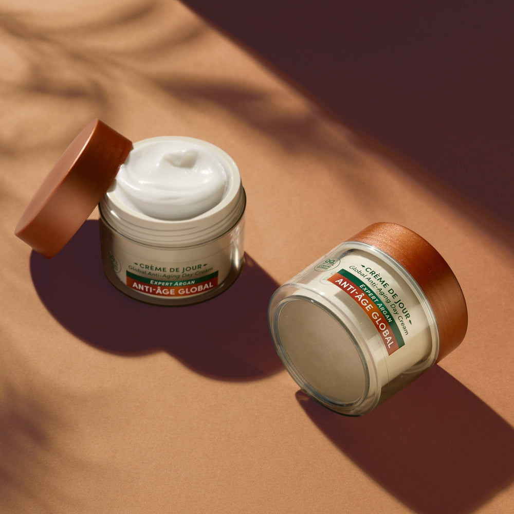 Deux pots de « Le Petit Olivier Duo Crème Jour et Baume Nuit Anti-âge », munis de couvercles en cuivre, reposent sur une surface marron. L'un des pots est ouvert, laissant entrevoir une crème blanche à l'intérieur. Les deux pots arborent des étiquettes vertes et rouges, idéales pour les soins hydratants du visage et les soins naturels.
