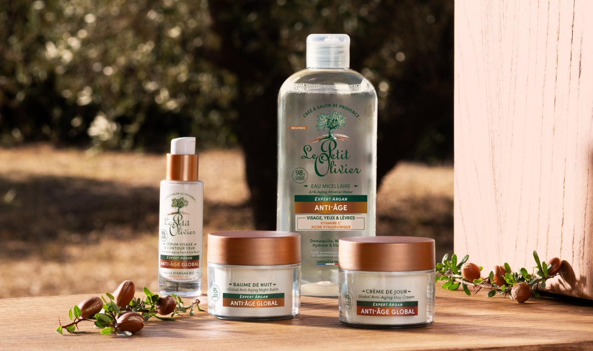 Quatre produits de soin « Le Petit Olivier », dont des crèmes et un sérum, sont présentés sur un support en bois à l'extérieur, avec des branches d'olivier et des oliviers en arrière-plan. Les produits portent la mention « Anti-Âge Global ».
