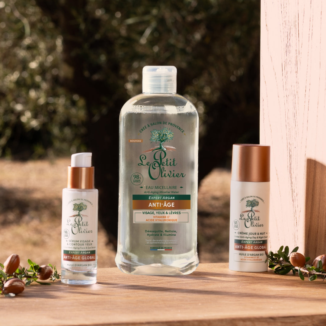 Trois produits « Le Petit Olivier Rituel Soin Visage Express Anti-Âge », présentés dans des emballages blancs et cuivrés, sont exposés à l'extérieur sur une surface en bois, entourés de feuilles vertes et de glands, sur un fond naturel flou.