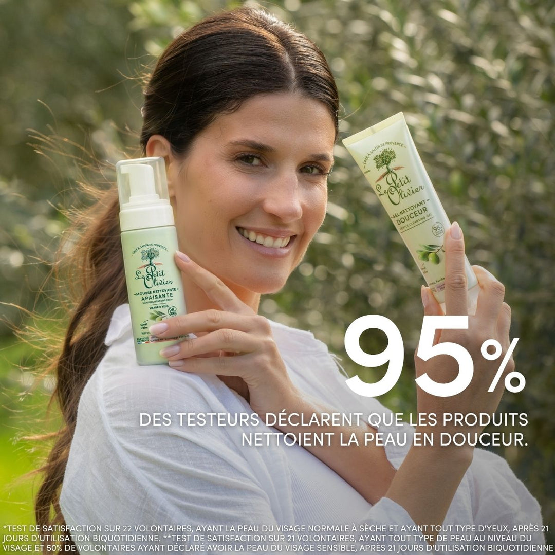 Une femme souriante, en extérieur, tient dans ses mains le « Duo Nettoyant Douceur - Jour & Nuit » de Le Petit Olivier, enrichi en ingrédients d'origine naturelle. 95 % des personnes ayant testé ce produit affirment qu'il nettoie la peau en douceur. Des arbres apparaissent à l'arrière-plan.