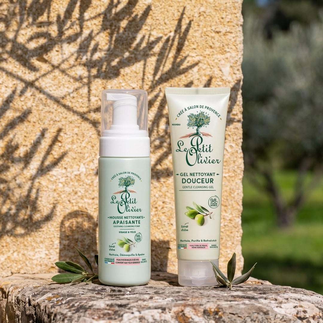 Le Duo Nettoyant Douceur - Jour & Nuit de Le Petit Olivier est présenté en extérieur sur une surface en pierre, entouré de feuilles d'olivier et avec, en arrière-plan, un jardin baigné de soleil, ce qui met en valeur ses ingrédients d'origine naturelle.