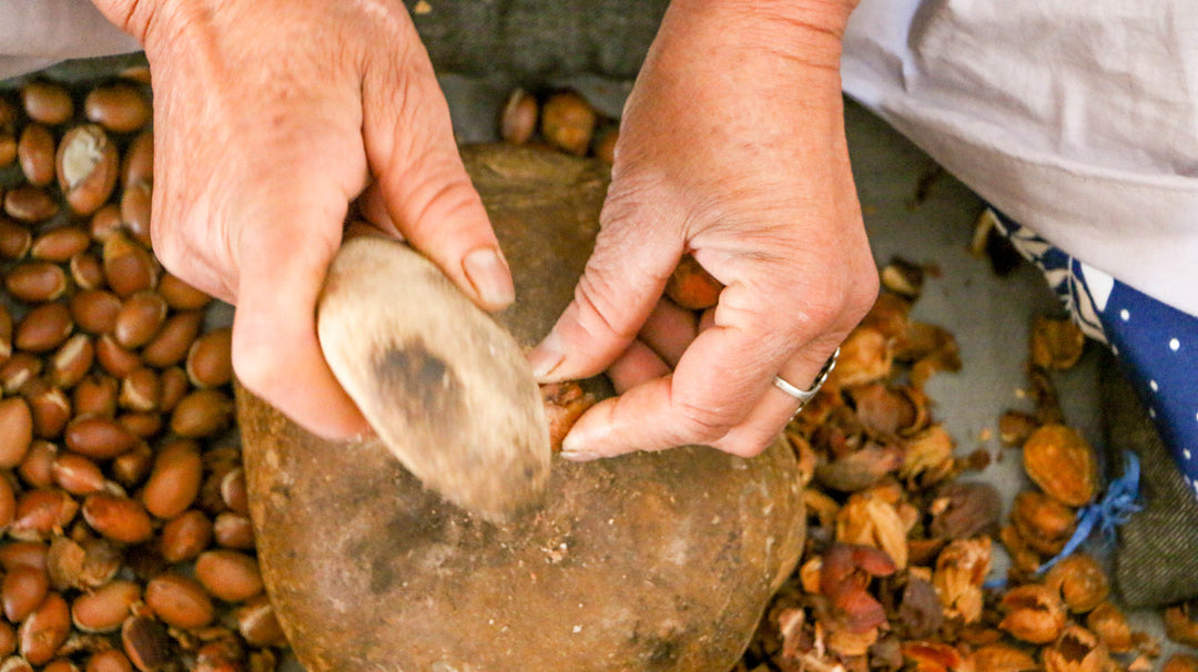 Deux mains utilisent un outil en pierre pour casser des noix d'argan, au milieu de coquilles éparpillées et de noix entières posées sur une surface plane.