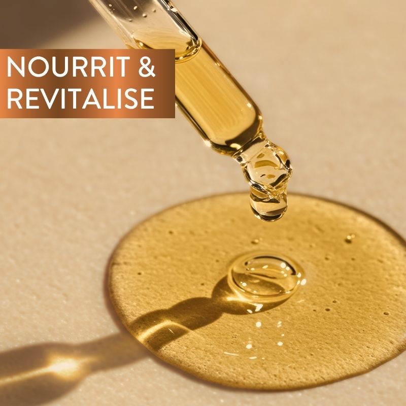 Un compte-gouttes déverse de l'huile dorée sur une surface lisse, formant une petite flaque. Les mots « NOURRIT & REVITALISE » apparaissent dans un encadré marron dans le coin supérieur gauche. Un éclairage chaleureux met en valeur la texture et l'éclat de l'huile.
