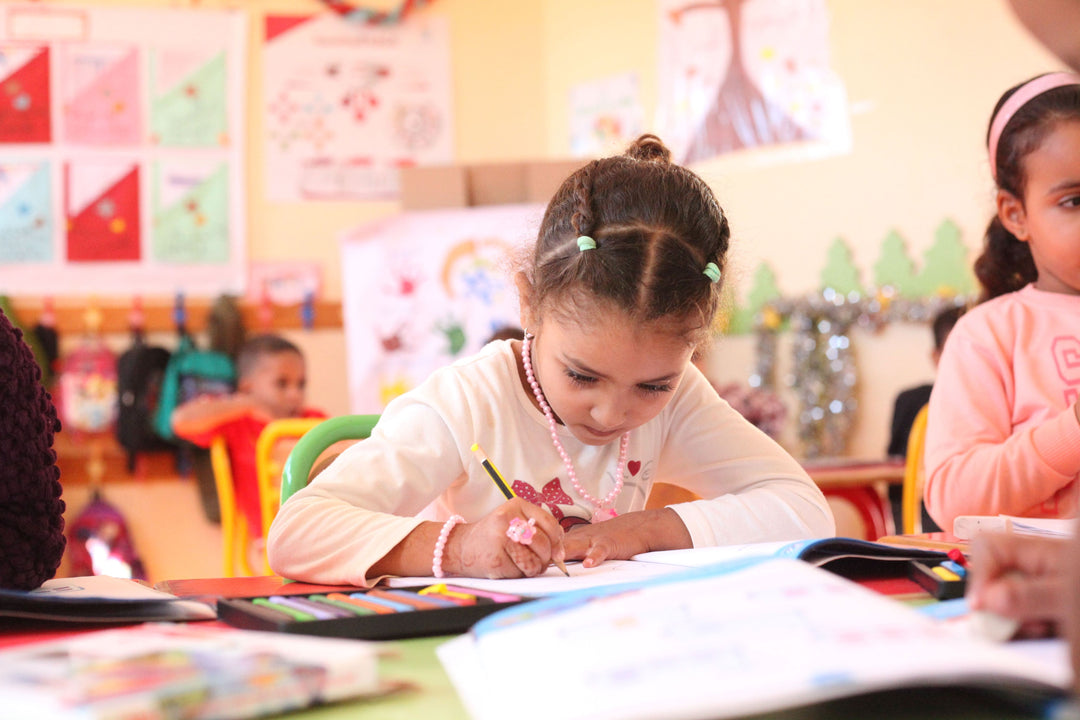 Une jeune fille aux cheveux tressés est assise à un pupitre, concentrée sur ce qu'elle écrit dans un cahier, entourée de fournitures scolaires colorées et d'autres enfants.