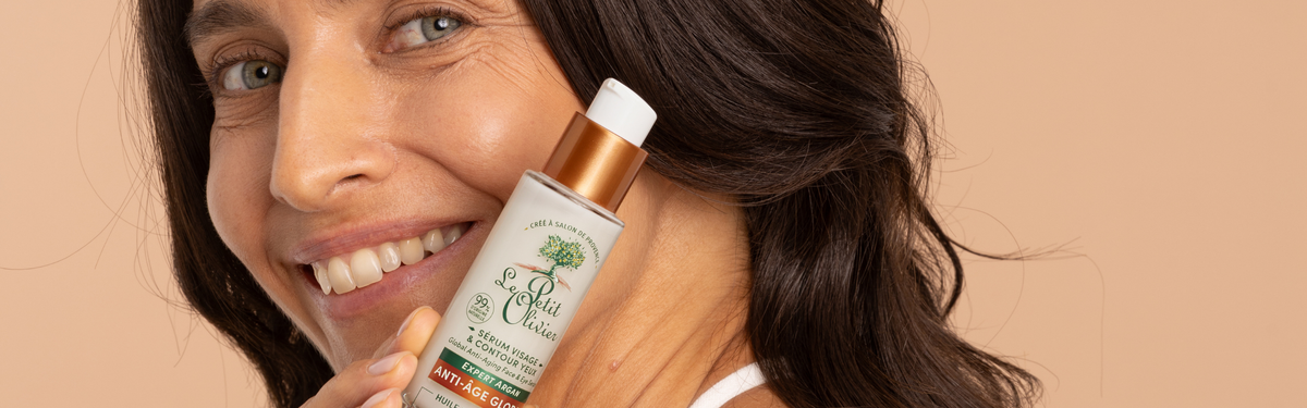 Une femme souriante aux longs cheveux foncés tient un flacon du produit de soin « La Petit Crème » près de son visage, sur un fond beige.