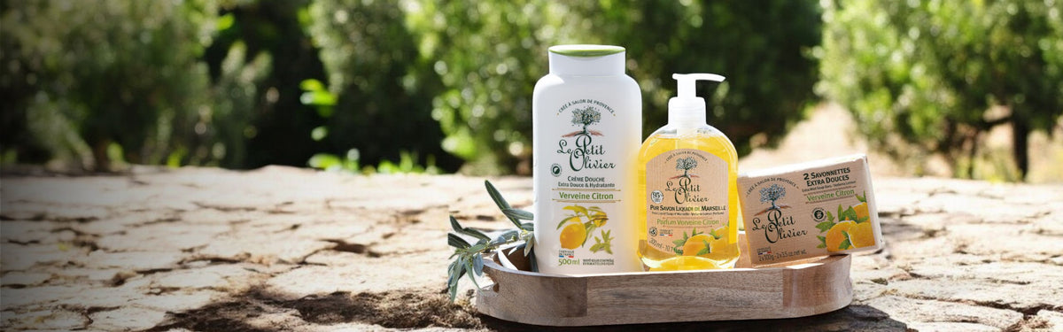 Sur une surface en pierre, un plateau en bois accueille le shampoing parfumé au citron « La Petit Olivier », du savon pour les mains et un savon en pain, avec des branches d'olivier à proximité et des arbres verts flous en arrière-plan.