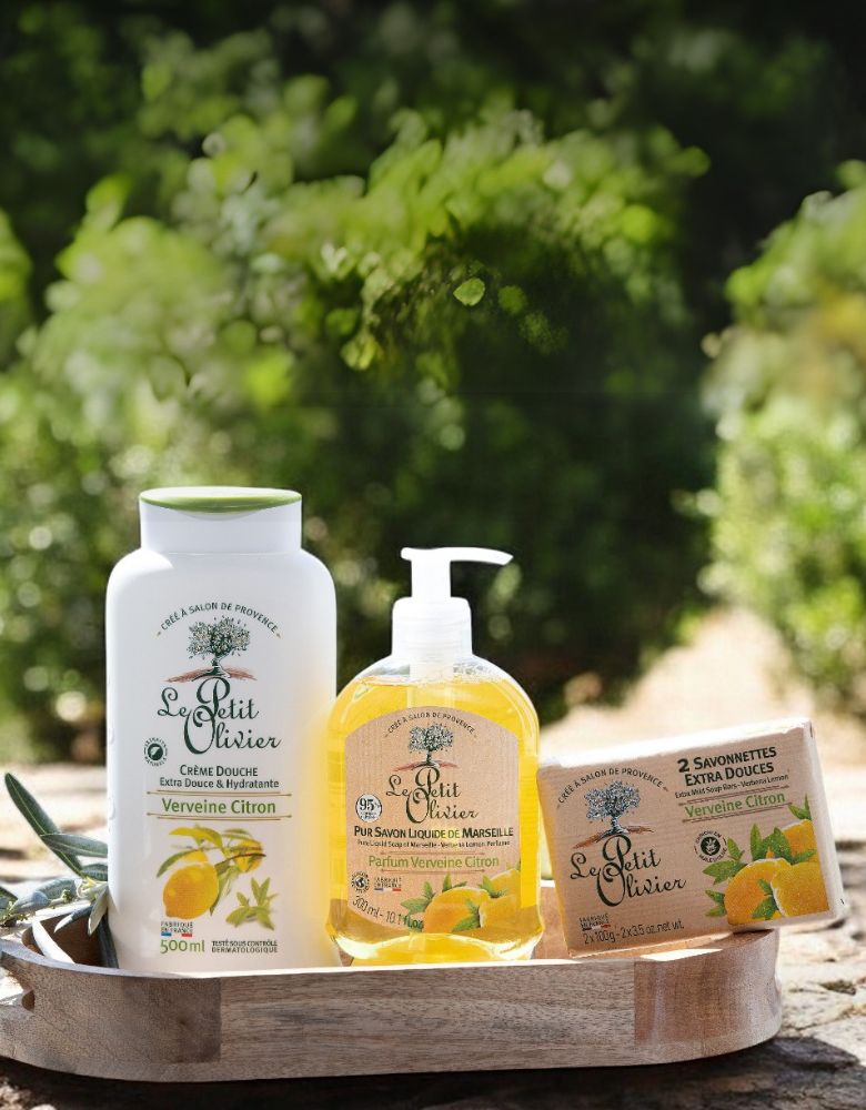 Trois produits de la gamme « Le Petit Olivier Verveine-Citron » — un flacon, un flacon-pompe et deux savons en pain — sont disposés sur un plateau en bois à l'extérieur, avec du feuillage vert et la lumière du soleil en arrière-plan.