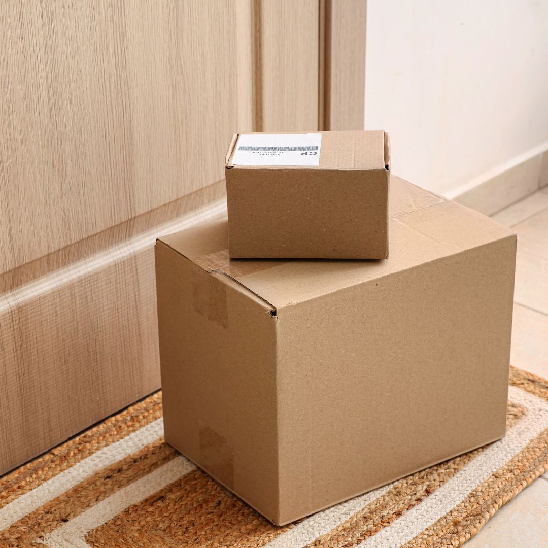 Deux colis en carton, un petit et un grand, sont posés sur un paillasson devant une porte en bois fermée, à l'intérieur d'une maison.