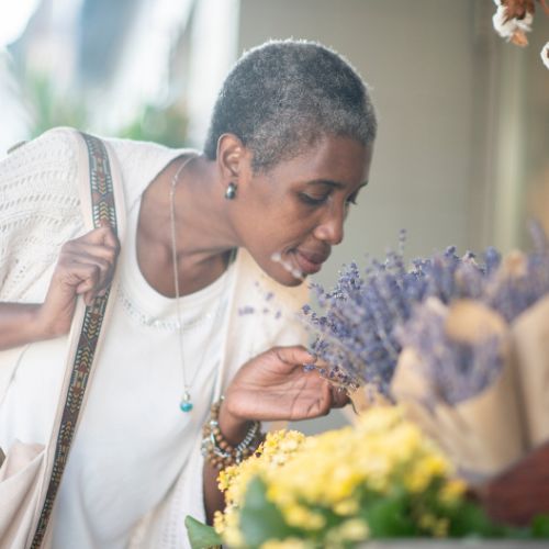 Une femme aux cheveux courts se penche en avant pour humer un bouquet de fleurs lavande et jaunes ; elle tient la bandoulière de son sac et porte un haut blanc ainsi que des bijoux.