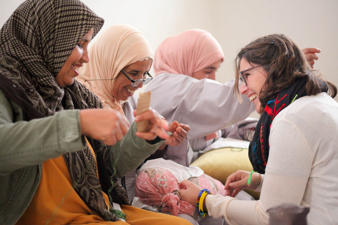 Quatre femmes sont assises ensemble, souriantes et rieuses, tandis qu'elles enveloppent un bébé dans une couverture à motifs roses. La scène dégage une atmosphère chaleureuse et joyeuse, suggérant une activité bienveillante ou collective autour du bébé.