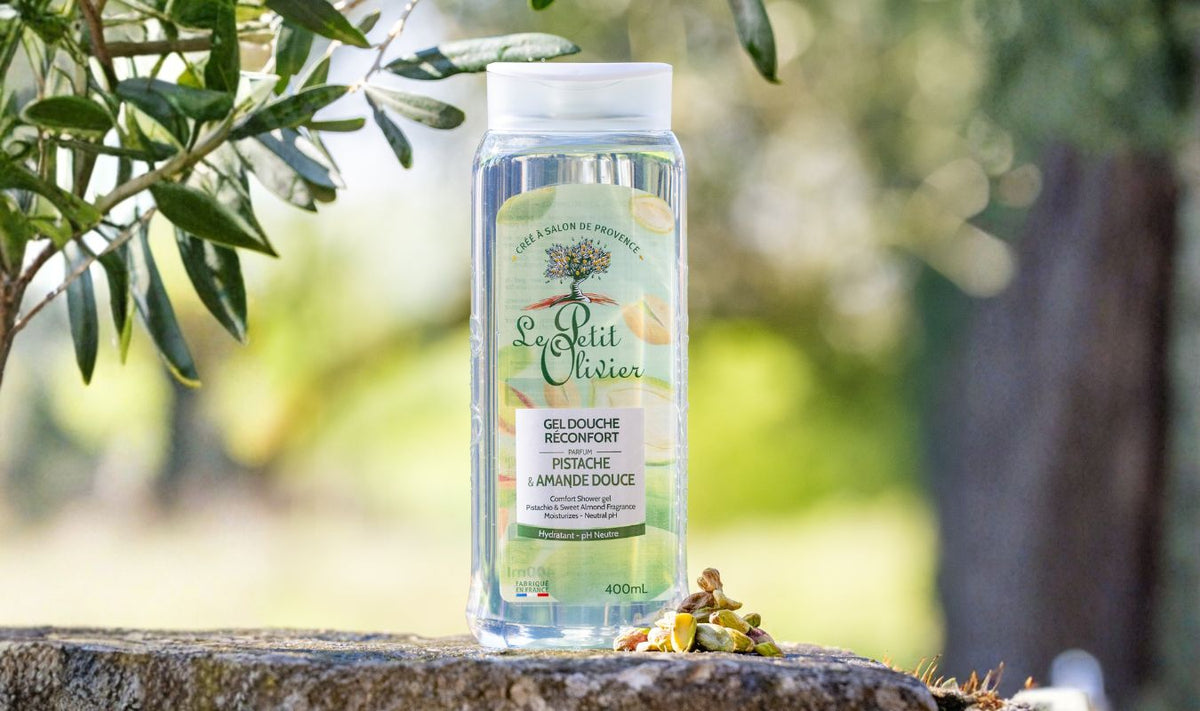 Un flacon de gel douche « Le Petit Olivier » est posé sur une surface en pierre à l'extérieur, entouré de pistaches et de feuilles, sur un fond vert naturel flou.
