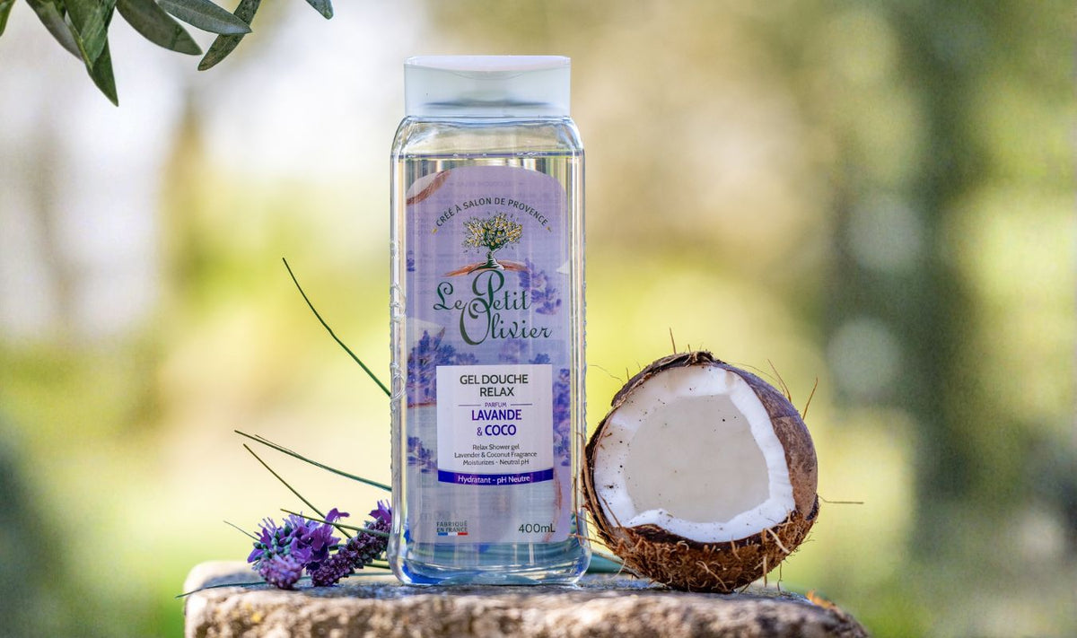 Un flacon de gel douche « Le Petit Olivier » à la lavande et à la noix de coco est posé sur une pierre, avec à côté une branche de lavande et une demi-noix de coco. L'arrière-plan est constitué d'une végétation floue.